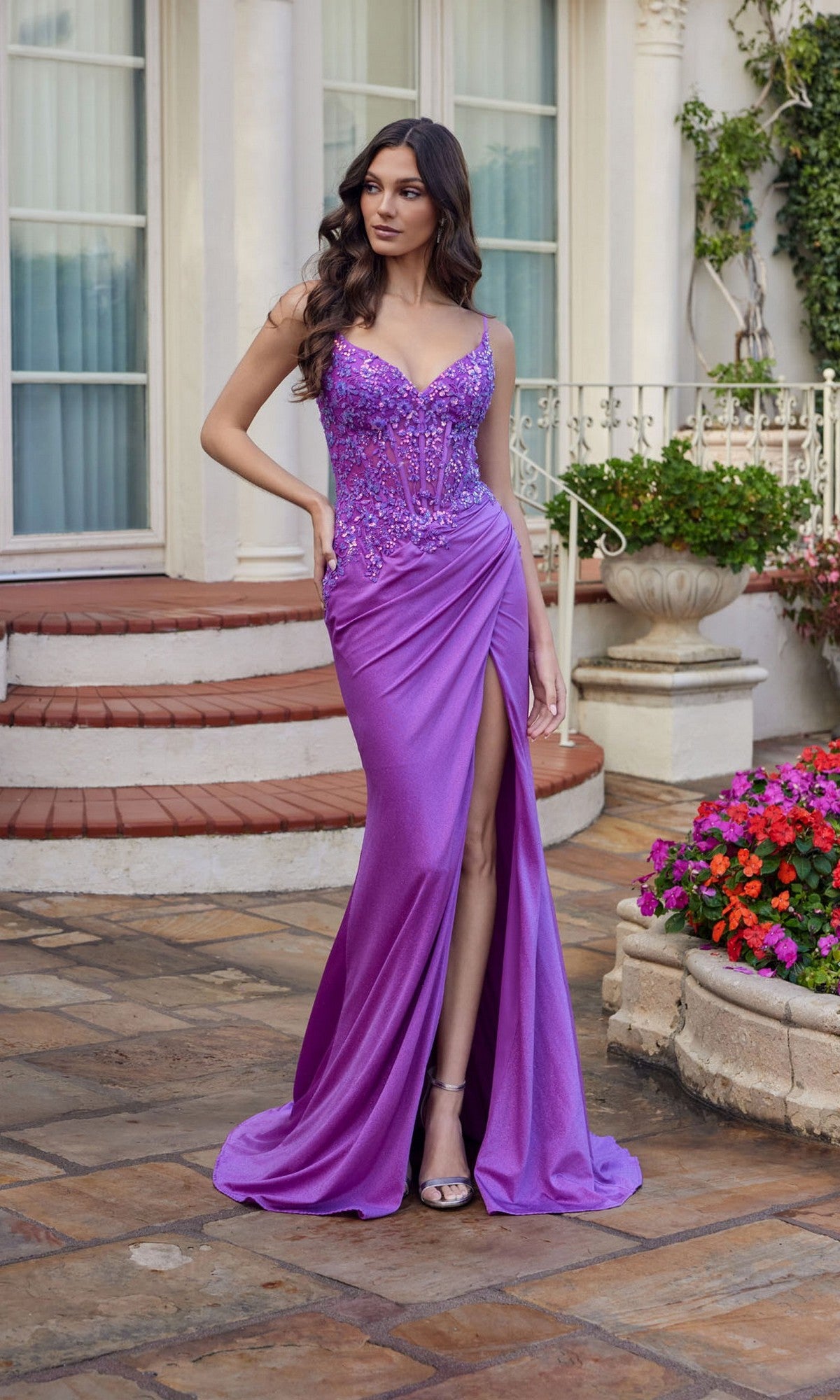 A woman wears the magenta Long Prom Dress: Juliet JT2613N, featuring a thigh-high slit and sheer corset, as she stands outdoors near stairs, potted plants, and a building with large windows.